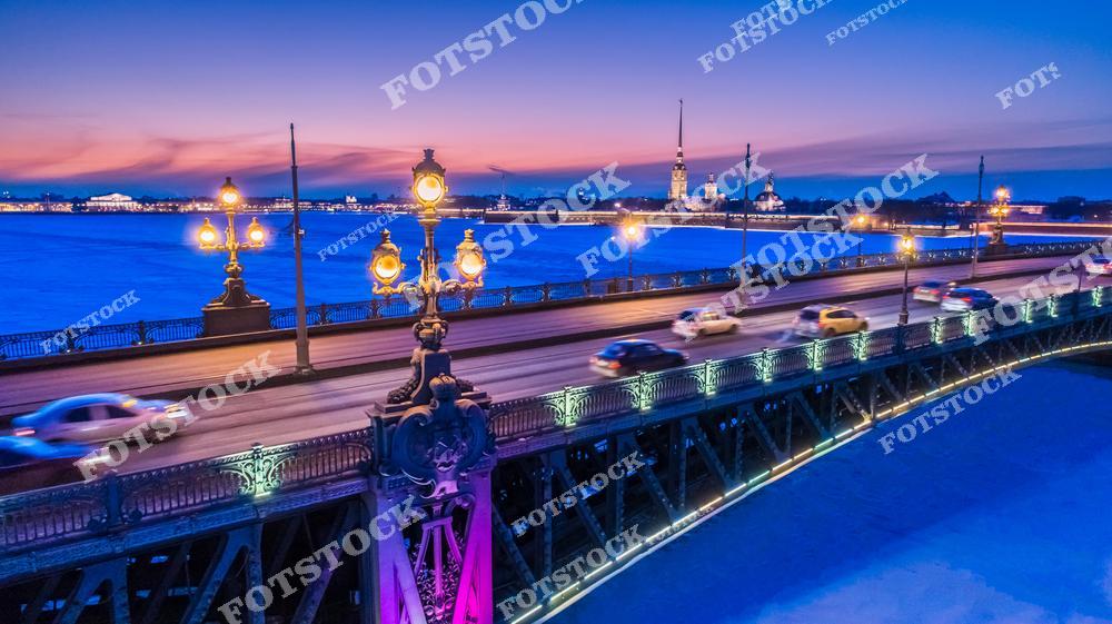 Фотография зимнего Санкт-Петербурга с панорамным видом с высоты, демонстрирующая Петропавловскую крепость, Троицкий мост, набережные Невы и историческую архитектуру города в вечернее время. Снимок показывает величественные мосты, замерзшие участки реки и огни города, создавая атмосферу спокойствия и романтики северной столицы России. Панорамный ракурс с дрона позволяет оценить масштабы улиц, набережных и городских сооружений, подчеркивая гармонию природы и архитектуры Петербурга. Изображение идеально подходит для туристических публикаций, зимних коллекций фотографий, постеров, интерьерного оформления и презентаций о городской архитектуре.